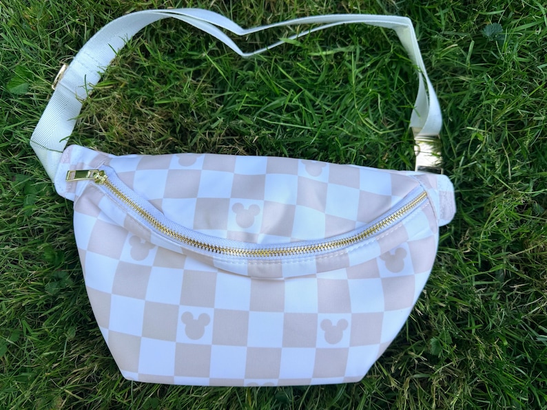 May include: A white and tan checkered fanny pack with a gold zipper and strap hardware. The bag features small Mickey Mouse silhouettes on the checkered pattern. The adjustable strap is white.