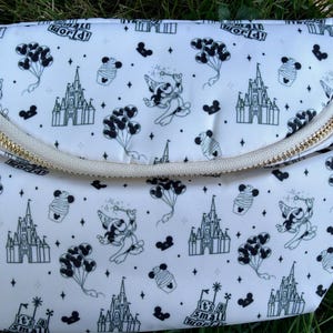May include: White fanny pack with a gold zipper and a repeating black and white pattern of a castle, Mickey Mouse, cupcakes, and balloons. The text "it's a small world!" is also printed on the bag.