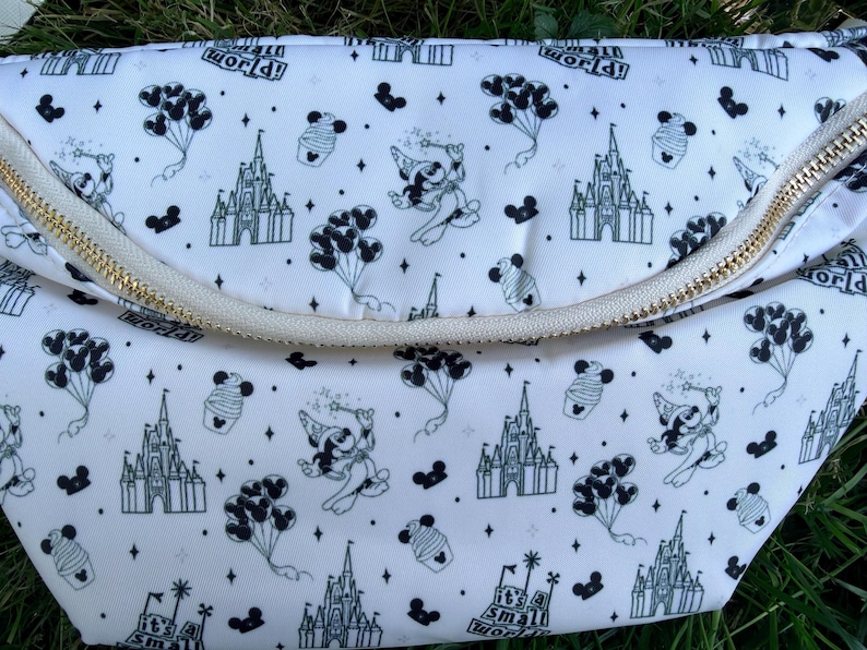 May include: White fanny pack with a gold zipper and a repeating black and white pattern of a castle, Mickey Mouse, cupcakes, and balloons. The text "it's a small world!" is also printed on the bag.