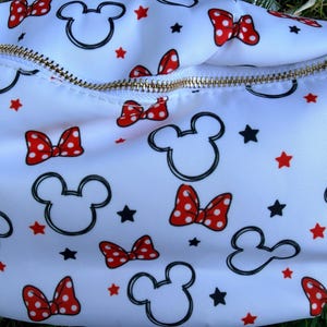 May include: White fanny pack with a gold zipper, featuring a pattern of Mickey Mouse heads, red polka dot bows, and black stars. The bag is made of a smooth, possibly nylon, material.