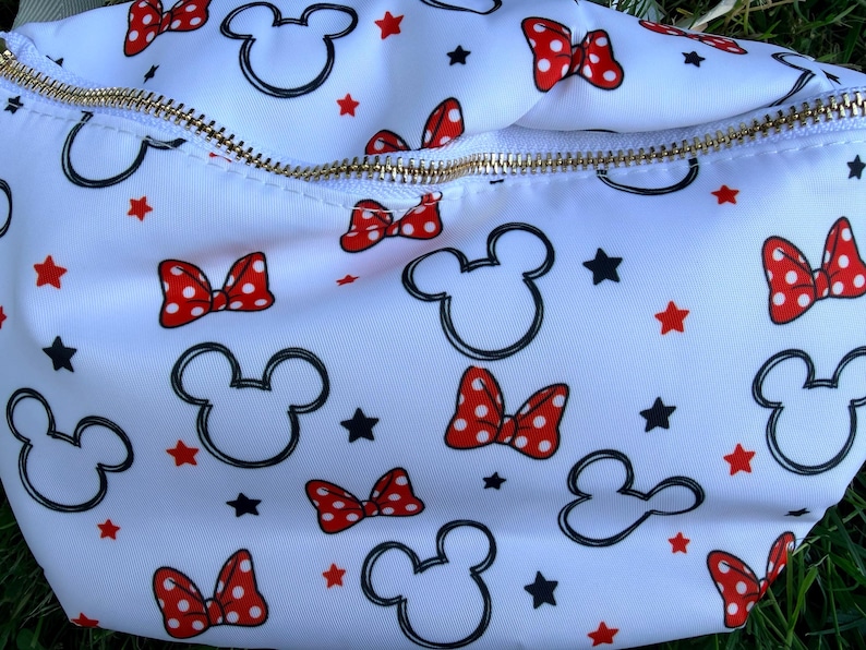 May include: White fanny pack with a gold zipper, featuring a pattern of Mickey Mouse heads, red polka dot bows, and black stars. The bag is made of a smooth, possibly nylon, material.