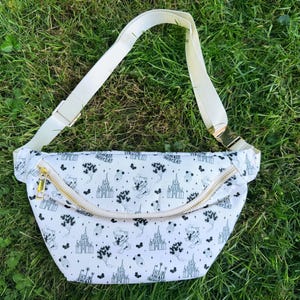 May include: White fanny pack with a black and white pattern featuring castles and cartoon characters. The bag has a cream-colored adjustable strap and a gold zipper. The bag is lying on a bed of green grass.