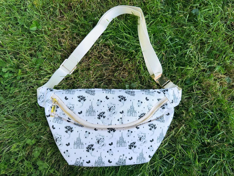 May include: White fanny pack with a black and white pattern featuring castles and cartoon characters. The bag has a cream-colored adjustable strap and a gold zipper. The bag is lying on a bed of green grass.