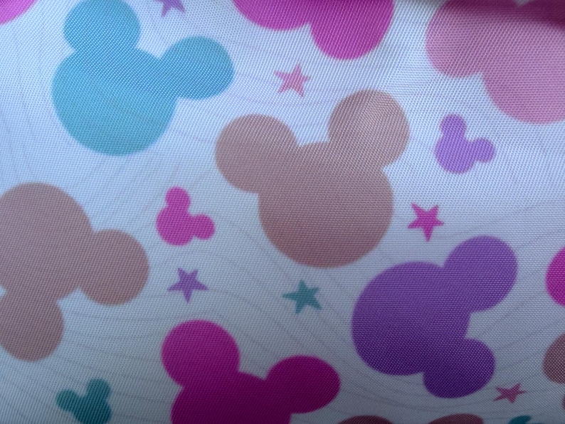 May include: A white fabric with a repeating pattern of Mickey Mouse silhouettes in various colors, including pink, purple, blue, and peach. Small star shapes are scattered throughout the design. The fabric has a subtle, wavy texture.