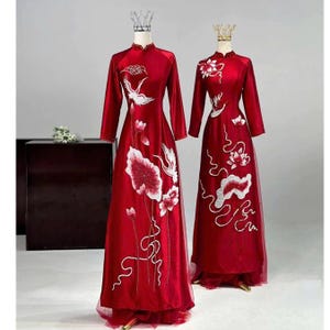 Puede incluir: Dos vestidos rojos de manga larga con cuellos mandarín y bordados blancos intrincados de flores y pájaros. Los vestidos se exhiben en maniquíes, mostrando sus siluetas fluidas y detalles delicados. Cada vestido tiene una falda transparente en capas.