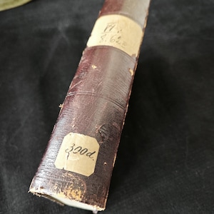 Puede incluir: Un libro antiguo cilíndrico con una cubierta de cuero marrón oscuro desgastada. El lomo tiene una etiqueta de papel de color claro con texto manuscrito. También es visible una etiqueta más pequeña con el texto "300d". El libro muestra signos de edad y desgaste.