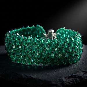 May include: A wide bracelet constructed from numerous faceted emerald green crystal beads arranged in a dense, interconnected lattice pattern, featuring a metallic silver-toned clasp mechanism visible at the back of the circular band.