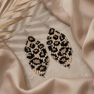 May include: A pair of dangle earrings featuring a leaf-shaped design crafted from small beads in a leopard print pattern. The beads are coloured in black, cream, and metallic gold, and the earrings are attached to silver-toned metal fishhook wires.