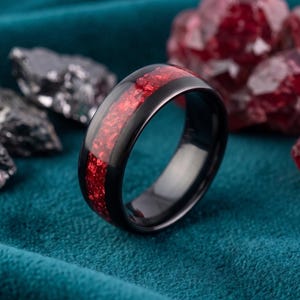 Puede incluir: Un anillo negro con una incrustación roja vibrante. El anillo se presenta sobre un fondo verde azulado con piedras grises oscuras y rojas. La banda del anillo es negra pulida y la incrustación roja tiene una apariencia texturizada.