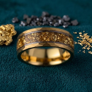 Puede incluir: Un anillo dorado con textura martillada y una banda central llena de escamas de oro. El anillo se muestra sobre un fondo verde azulado oscuro, con pepitas de oro y piedras grises oscuras.