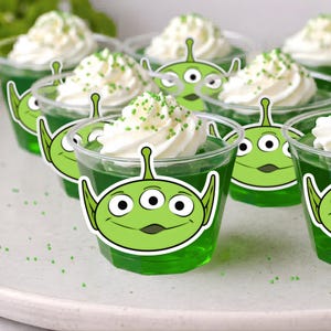 May include: A tray of green gelatin desserts topped with whipped cream and green sprinkles. Each dessert is in a clear plastic cup and decorated with a green alien face sticker. A fun and festive treat.