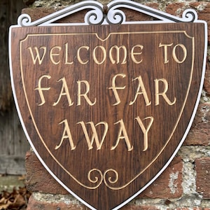 以下が含まれることがあります： 「WELCOME TO FAR FAR AWAY」の文字が彫られた、盾型の木製サイン。白い縁取りと上部の装飾的な金属細工が特徴です。レンガの壁に取り付けられています。
