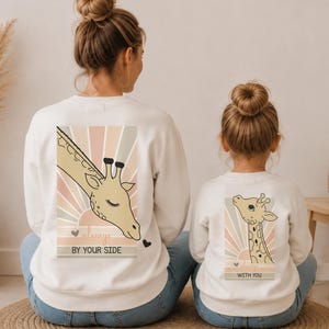 May include: Two white crewneck sweatshirts featuring a graphic of a giraffe head with closed eyes against a sunburst background. The adult sweatshirt displays the text "always BY YOUR SIDE" and the child sweatshirt displays "ALWAYS WITH YOU".