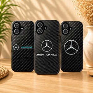 Puede incluir: Tres fundas negras para teléfono con textura de fibra de carbono. Cada funda presenta un logotipo diferente de Mercedes-Benz AMG. La funda de la izquierda tiene un logotipo y texto turquesa, la funda del medio tiene un logotipo plateado y la palabra "AMG", y la funda de la derecha tiene un logotipo plateado.