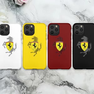Può includere: Quattro custodie per telefono bianche, gialle, rosse e nere. Ogni custodia presenta il logo Ferrari: un cavallo nero rampante su uno scudo giallo. Le custodie sono esposte su una superficie marmorizzata.
