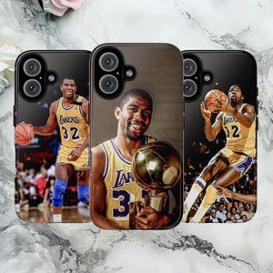 May include: Three phone cases featuring basketball player Magic Johnson in a Lakers uniform. The cases display images of Johnson holding a basketball and a trophy. The cases are black with a brown background.