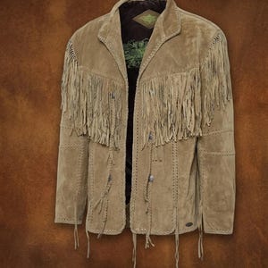 Könnte beinhalten: Hellbraune Wildlederjacke mit langen Fransen an Ärmeln und Brust. Die Jacke hat einen Reverskragen, Ziernähte und silberfarbene Knöpfe. Das Innenfutter ist dunkelbraun.