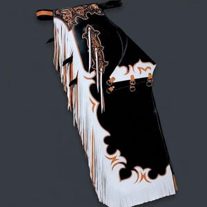 Peut inclure: Chaps en cuir noir avec franges blanches et détails décoratifs orange et dorés. Les chaps présentent des motifs floraux et de volutes complexes, avec des sangles à boucles et une ceinture. Vêtement de style western.
