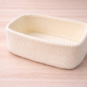 May include: A rectangular, cream-coloured crochet basket. The basket has a rounded shape and is made of a textured, knitted material. The basket is empty and sits on a light-coloured wooden surface.