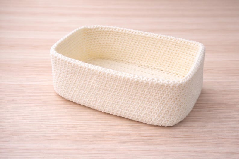 May include: A rectangular, cream-coloured crochet basket. The basket has a rounded shape and is made of a textured, knitted material. The basket is empty and sits on a light-coloured wooden surface.