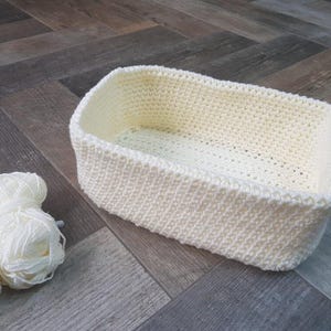 May include: A cream-coloured, rectangular crochet basket with rounded corners is on a wooden surface. Three balls of white yarn are next to the basket. The basket is made of thick, textured yarn, and is likely handmade.