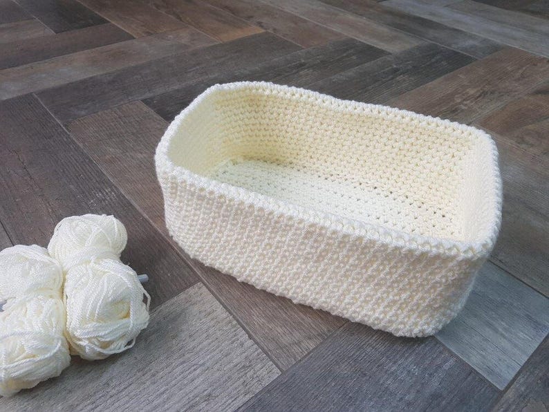May include: A cream-coloured, rectangular crochet basket with rounded corners is on a wooden surface. Three balls of white yarn are next to the basket. The basket is made of thick, textured yarn, and is likely handmade.