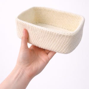 May include: A rectangular, off-white crochet basket with a textured stitch pattern. The basket is held in a hand, showcasing its handmade quality. The neutral color and simple design suggest a versatile storage solution.