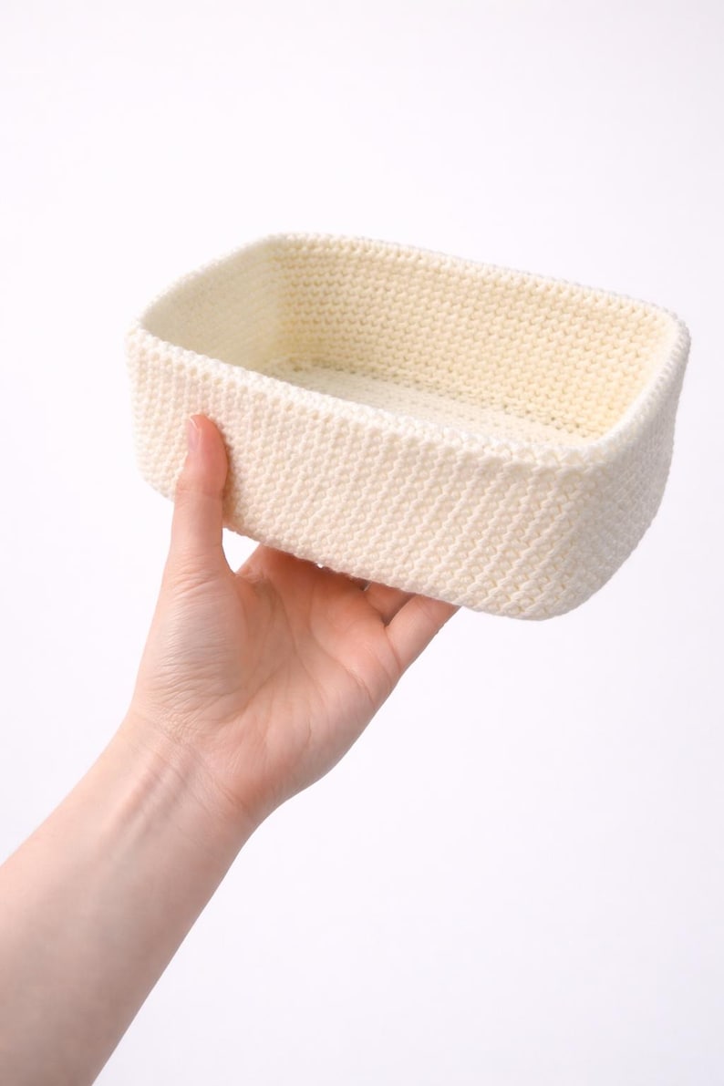 May include: A rectangular, cream-coloured crochet basket with a textured stitch pattern. The basket is held in a hand, highlighting its handmade quality. The neutral colour and simple design suggest a versatile storage solution.