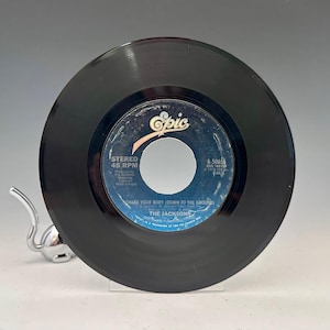 May include: A black vinyl record with a blue and white label. The label reads "Shake Your Body (Down to the Ground)" by The Jacksons. The record is a 45 RPM single.