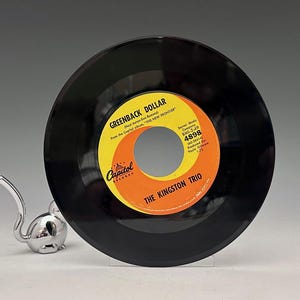 Puede incluir: Un disco de vinilo negro con una etiqueta amarilla y naranja. La etiqueta dice "GREENBACK DOLLAR" de The Kingston Trio. El logotipo de Capitol Records es visible. Una figura de ratón plateado está en primer plano.