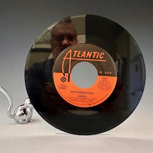 May include: A black vinyl record with an orange and white label that reads "Misunderstanding" by Genesis. The record is 7 inches in diameter. A silver mouse figurine is in the foreground.