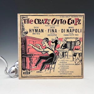 May include: Vintage record album cover for "The Crazy Otto Cafe" featuring Dick Hyman, Jack Fina, and Mike Di Napoli. The cover art depicts a cartoon scene of a pianist, musicians, and patrons in a cafe setting. The album is printed in the USA.