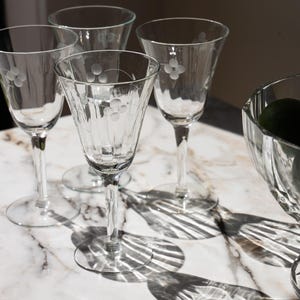May include: A set of four clear glass stemmed wine glasses featuring a delicate etched floral pattern on the bowl and vertical fluting along the lower section. Each glass has a slender stem and a circular base, reflecting light onto the surface.