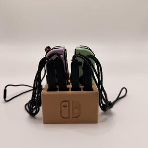 Könnte beinhalten: Eine braune Ladestation für eine Spielkonsole, die mehrere farbige Controller hält. Die Controller sind in Lila, Grün und Schwarz gehalten und haben schwarze Kabel. Auf der Dockingstation befindet sich das Logo der Konsole.