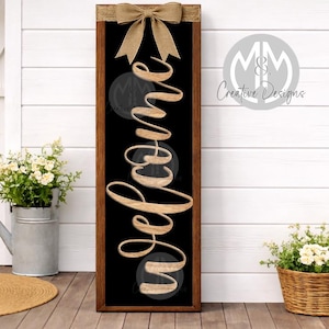 May include: A rectangular wooden welcome sign with a black background and the word "welcome" in a cursive font. The sign is adorned with a burlap bow at the top. The sign is displayed on a porch with plants and a welcome mat.