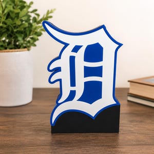 Könnte beinhalten: Ein freistehendes Holzschild in Form des Buchstabens D in einer gotischen Schriftart. Der Buchstabe ist in Königsblau mit einer dicken weißen Umrandung gehalten und steht auf einem soliden, rechteckigen schwarzen Sockel.