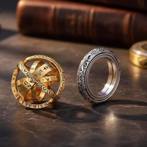 May include: Two rings on a dark surface. One is gold-colored, spherical, and has engraved bands with letters and symbols. The other is silver-colored with an ornate, engraved design around the band.