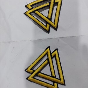 May include: Two embroidered patches, each featuring a yellow triangle design with a thick black border. The geometric pattern consists of two overlapping triangles, creating an interlocking effect with bright yellow thread and black stitching.