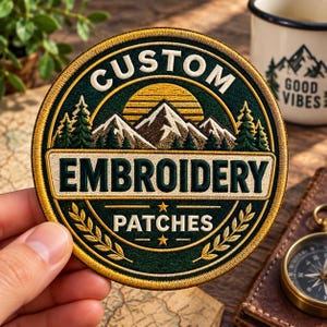 May include: A circular embroidered patch featuring a dark green background with a mountain range, pine trees, and a yellow sun. The text reads CUSTOM EMBROIDERY PATCHES in cream-colored lettering. The patch has a gold-colored stitched border.