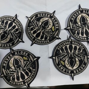 May include: Five circular embroidered patches featuring a black and white design of a mountain landscape, a moon, and a compass needle. The text NEVER ALONE FURTHER TOGETHER is stitched around the border with the letters BH in yellow.