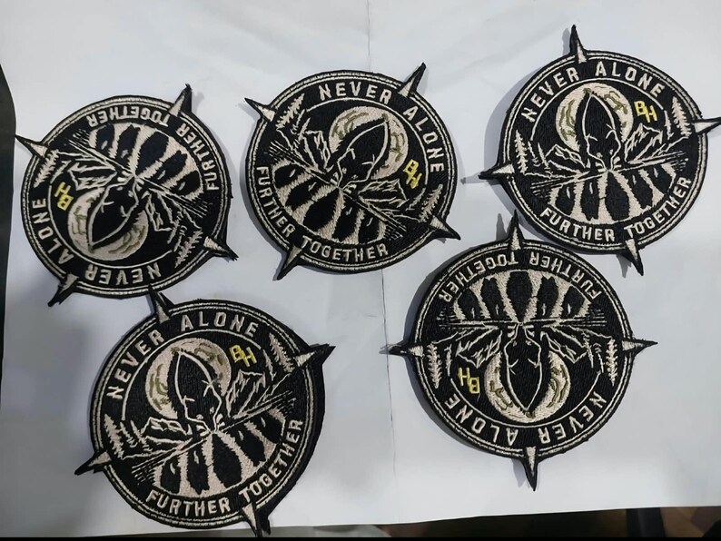 May include: Five circular embroidered patches featuring a black and white design of a mountain landscape, a moon, and a compass needle. The text NEVER ALONE FURTHER TOGETHER is stitched around the border with the letters BH in yellow.