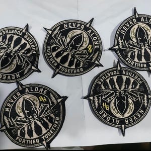 May include: Five circular embroidered patches featuring a black and white design of a mountain landscape, a moon, and a compass needle. The text "NEVER ALONE" and "FURTHER TOGETHER" is stitched along the border, with the letters "BH" in yellow.