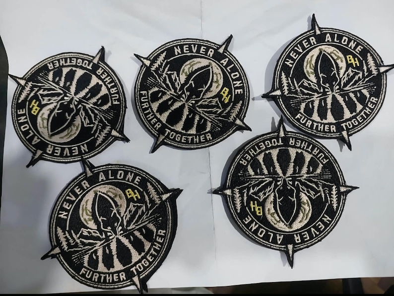 May include: Five circular embroidered patches featuring a black and white design of a mountain landscape, a moon, and a compass needle. The text "NEVER ALONE" and "FURTHER TOGETHER" is stitched along the border, with the letters "BH" in yellow.