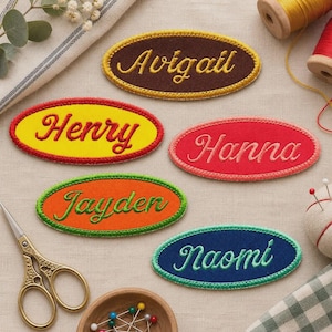 May include: Five oval-shaped embroidered fabric patches featuring the names Avigail, Henry, Hanna, Jayden, and Naomi. Each patch has a distinct background color including brown, yellow, red, orange, and blue, with contrasting thread for the names.