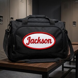 Puede incluir: Una bolsa de deporte negra con un parche ovalado central de fondo blanco y borde rojo. El nombre Jackson est&aacute; bordado en rojo con letra cursiva en el centro del &oacute;valo. La bolsa incluye un asa superior, una correa para el hombro y bolsillos laterales de malla.