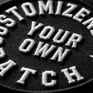 K&ouml;nnte beinhalten: Ein runder, schwarzer, bestickter Aufn&auml;her mit wei&szlig;em, gesticktem Text, der CUSTOMIZE YOUR OWN PATCH in einer fetten serifenlosen Schrift zeigt. Die Stickerei hat eine erhabene, strukturierte Optik auf dem dunklen Stoff.