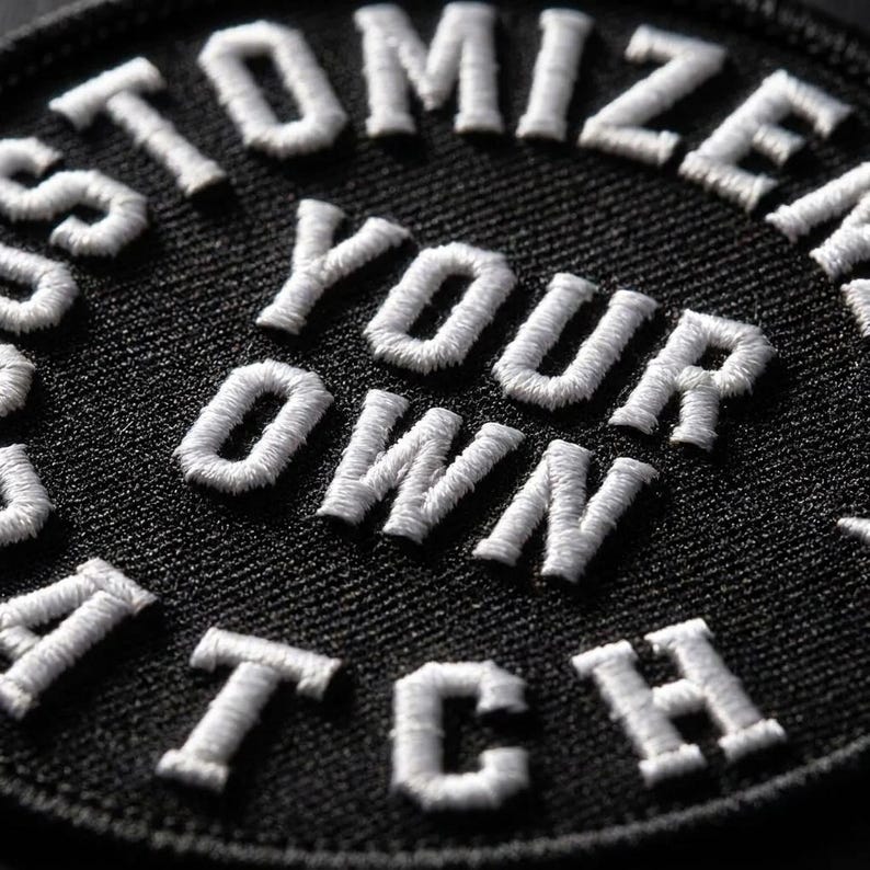 K&ouml;nnte beinhalten: Ein runder, schwarzer, bestickter Aufn&auml;her mit wei&szlig;em, gesticktem Text, der CUSTOMIZE YOUR OWN PATCH in einer fetten serifenlosen Schrift zeigt. Die Stickerei hat eine erhabene, strukturierte Optik auf dem dunklen Stoff.