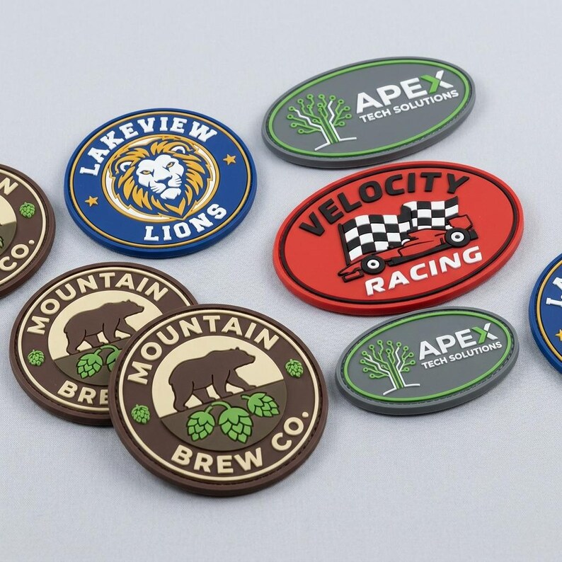 May include: A collection of circular and oval rubber patches featuring various designs including the Lakeview Lions logo, a red Velocity Racing emblem with a checkered flag, Apex Tech Solutions branding, and Mountain Brew Co. logos with bear graphics.
