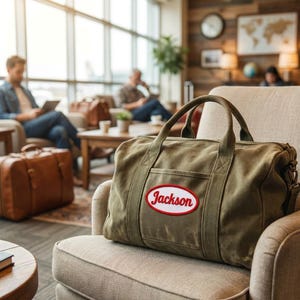 Puede incluir: Una bolsa de viaje de lona verde oliva con dos asas superiores y un bolsillo lateral. La bolsa presenta un parche ovalado blanco con borde rojo que contiene el texto cursivo rojo Jackson. La bolsa descansa sobre una silla beige tapizada.