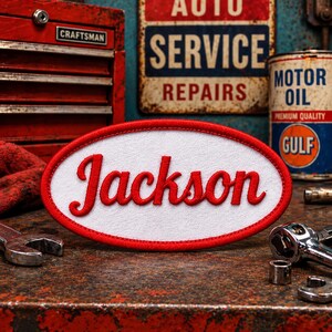 May include: An oval-shaped embroidered patch featuring a white background with a red border and the name Jackson stitched in red cursive script. The patch is designed with a clean, bold aesthetic, showcasing the name in a prominent, stylized font.